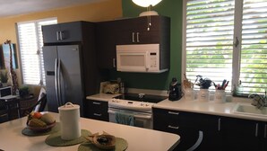 A compact kitchen with a refrigerator, microwave, stove, and sink. There is a dining table with a bowl of fruit and a pitcher.