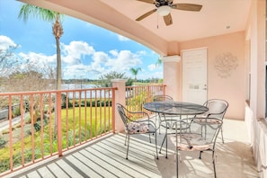 A balcony with a table and chairs, a ceiling fan, and a view of a landscaped area with trees and a body of water.
