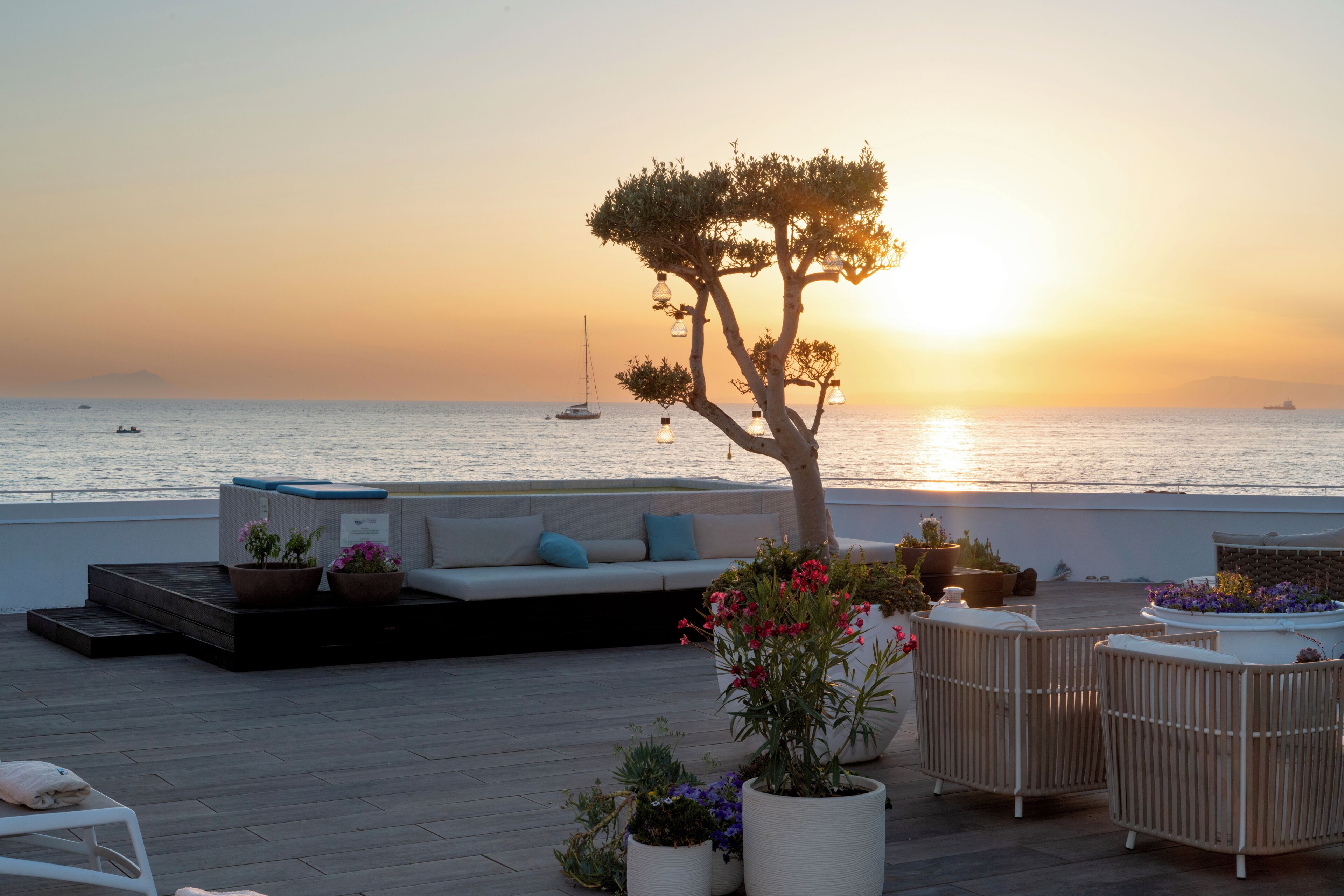 Una terraza en la azotea con sofá, sillas, plantas en macetas y vistas al mar al atardecer.