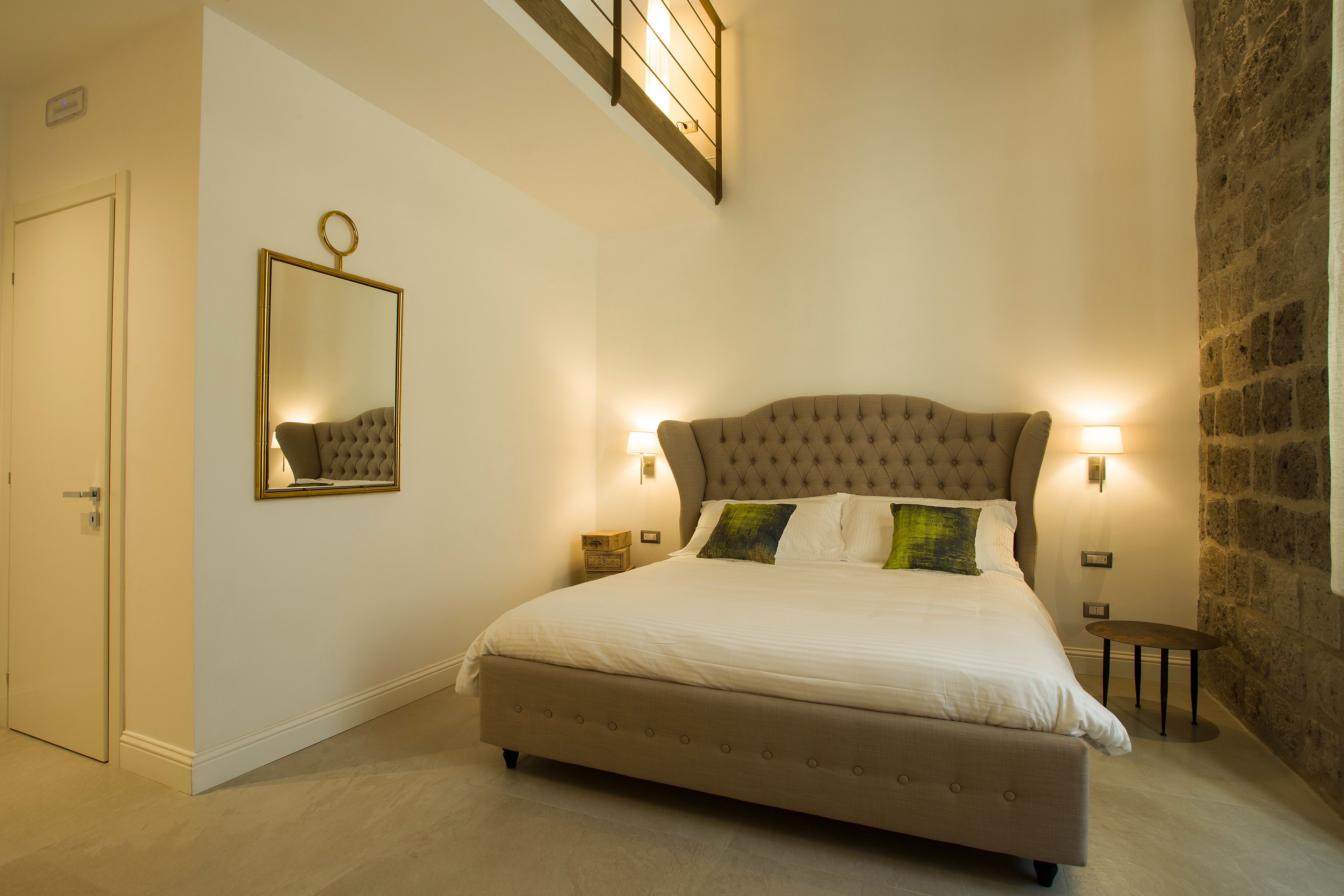 A bedroom with a large bed, a mirror, two bedside tables, a wall-mounted light, and a stone wall.