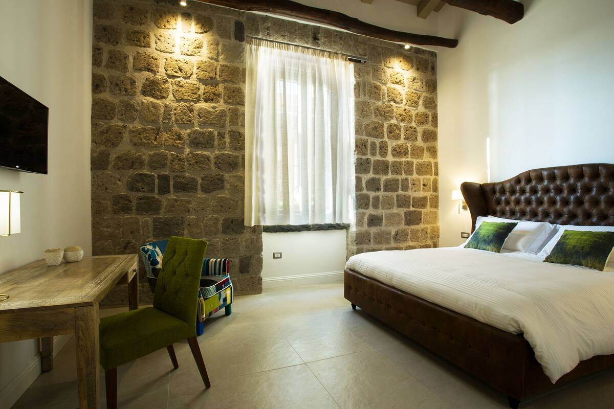 Un dormitorio con pared de piedra, una cama con cabecera marrón, una silla verde y una mesa de madera.