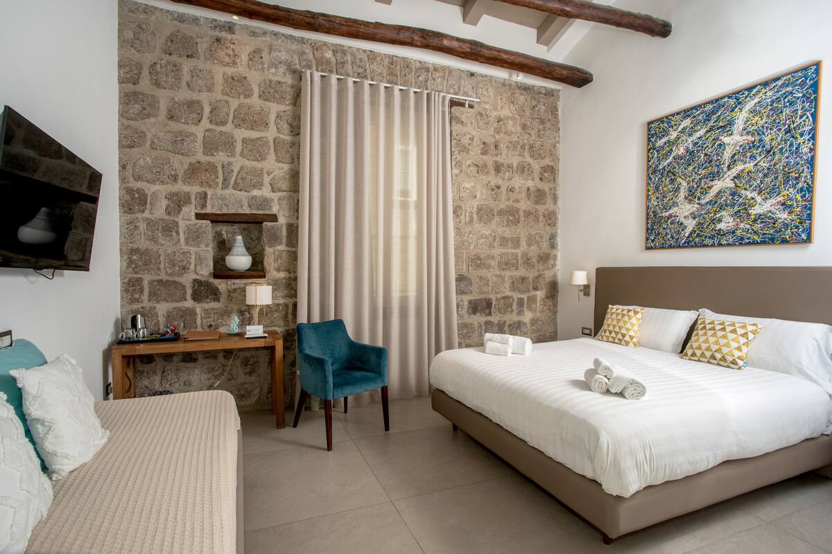 Un dormitorio con pared de piedra, una cama con ropa de cama blanca, una silla azul y un cuadro en la pared.