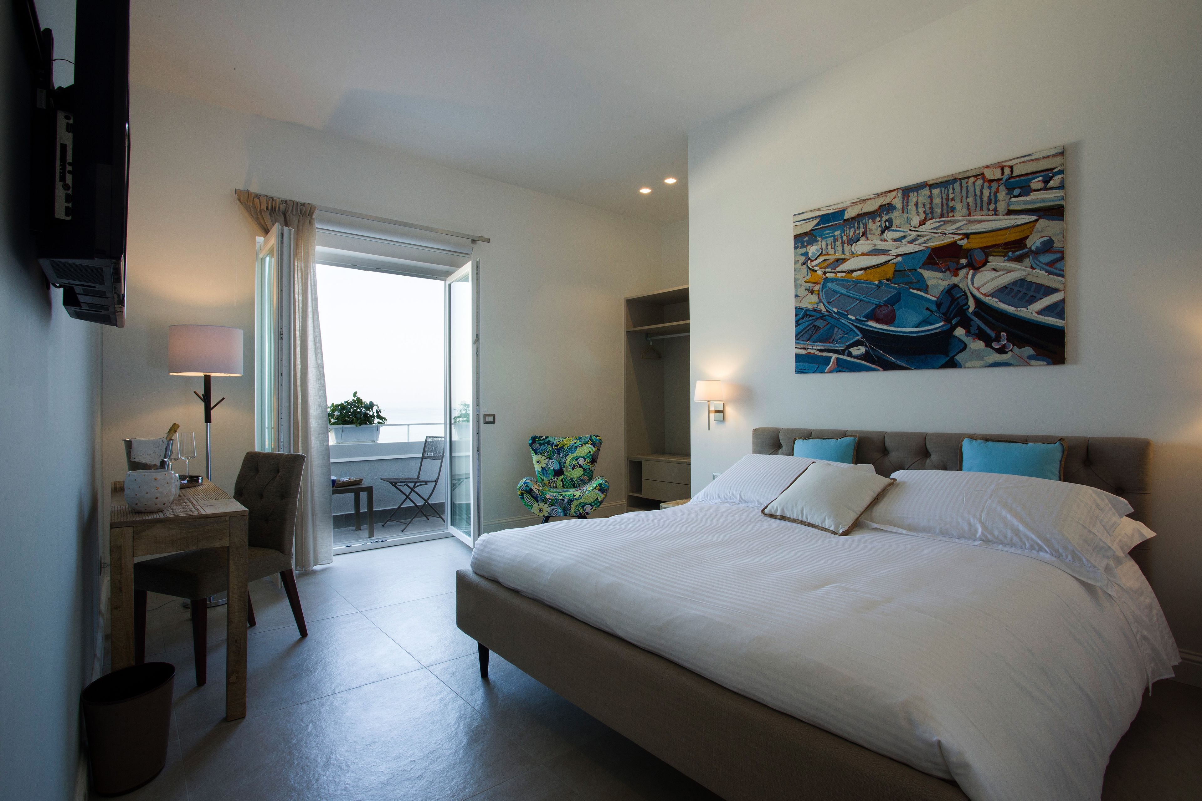 A modern hotel room with a large bed, a desk with a chair, a painting on the wall, and a view through a glass door.