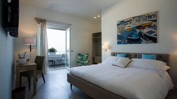 A modern hotel room with a large bed, a desk with a chair, a painting on the wall, and a view through a glass door.