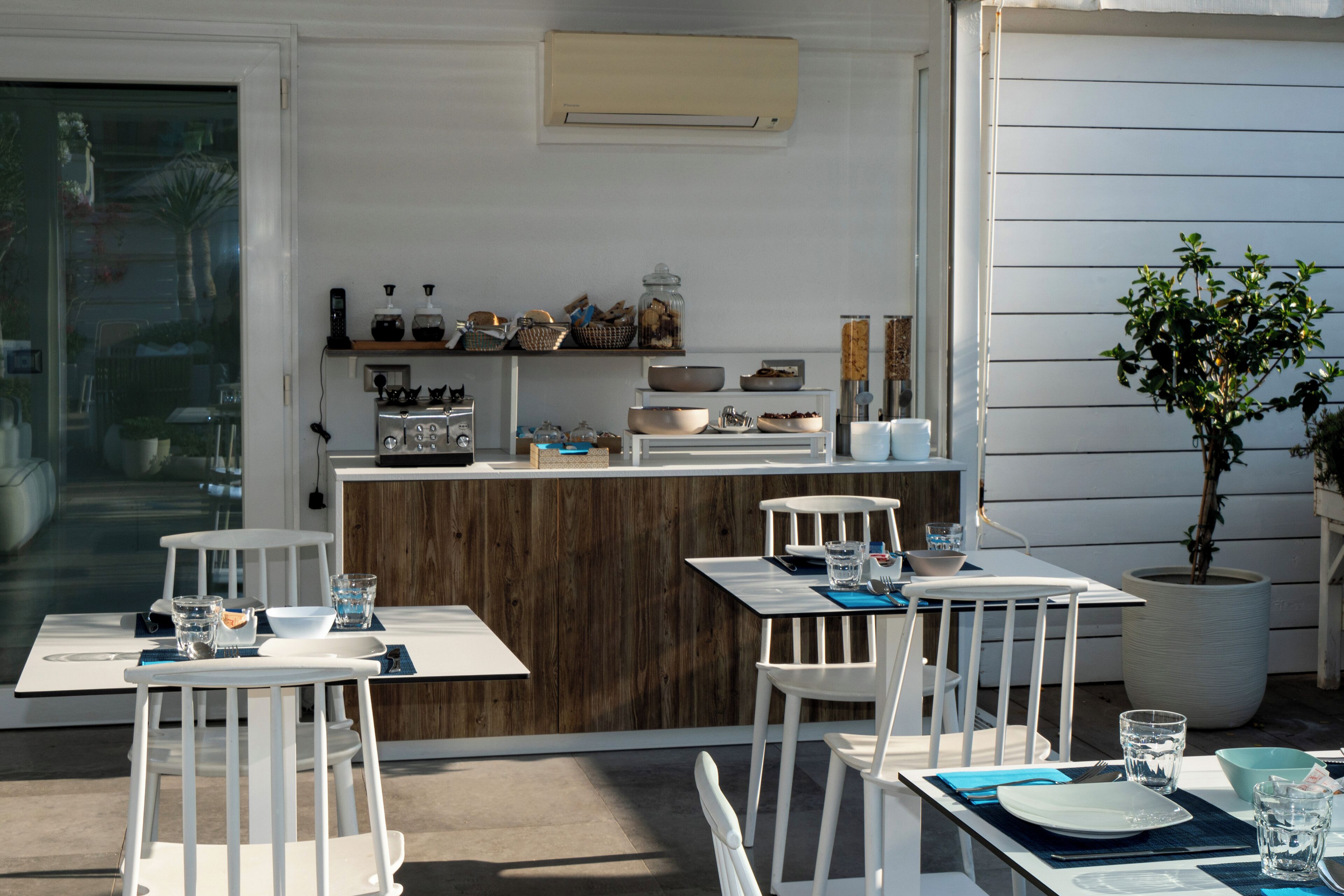 An outdoor dining area with white chairs and tables, a wooden counter with kitchenware, and a potted plant.