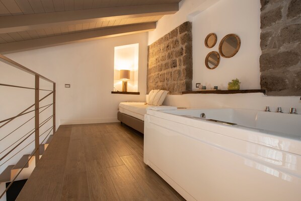 A modern bathroom with a large white bathtub, a wooden floor, a stone wall, and a small window with a lamp. - La Chiaja art&relais (Castellammare di Stabia)