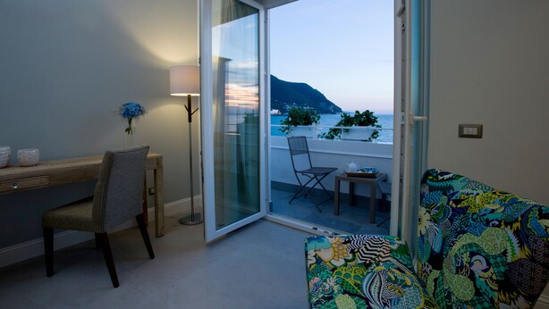 Ein Zimmer mit Schreibtisch, Stuhl und einem farbenfrohen Sofa. Eine Glastür führt zu einem Balkon mit Blick auf das Meer und die Berge.