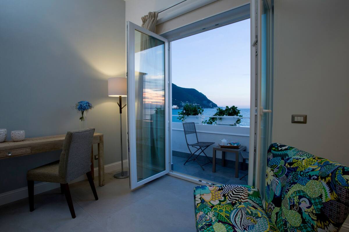 Una habitación con escritorio, silla y un sofá colorido. Hay una puerta corrediza de vidrio que da a un balcón con vistas al mar y las montañas.