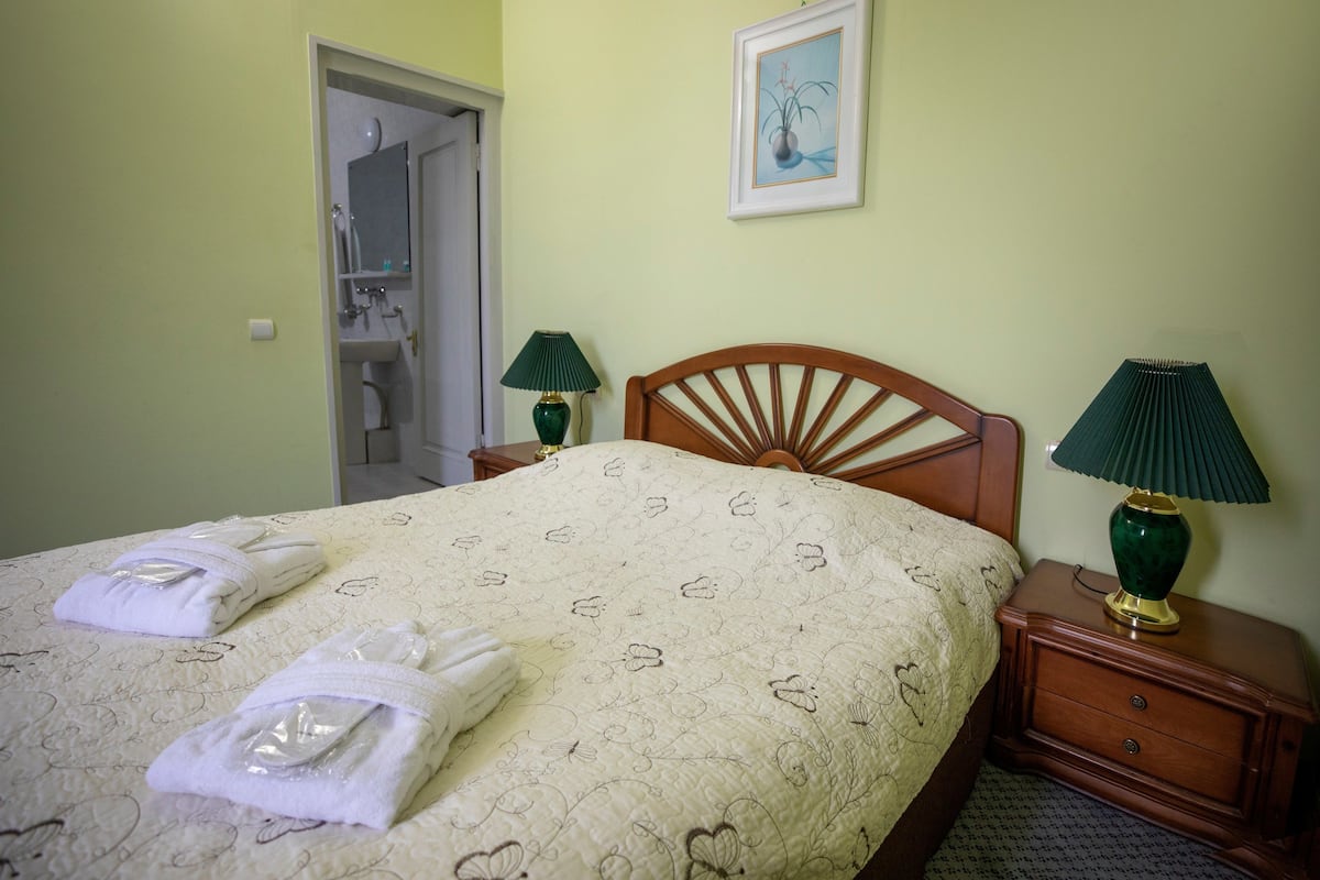 A neatly made bed with a floral-patterned bedspread and two folded towels. There are two bedside tables with lamps, a wooden headboard, and a framed picture on the wall.