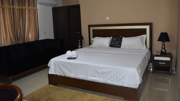 A neatly arranged hotel room with a large bed, a bedside table with a lamp, a dark-colored sofa, and a small rug. - Cascadehotel (Accra)