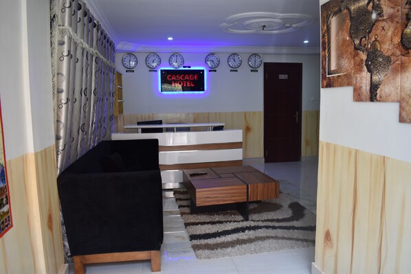 A hotel lobby with a black sofa, wooden coffee table, and a TV displaying "CASCADE HOTEL". - Cascadehotel (Accra)