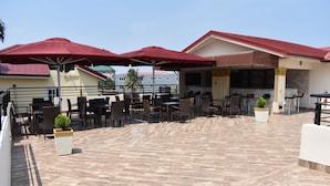 An outdoor patio with multiple tables and chairs, red umbrellas, and a barbecue area. - Cascadehotel (Accra)