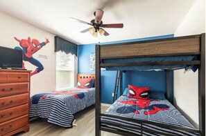 Een slaapkamer met twee stapelbedden, elk met een Spider-Man dekbedovertrek. Er staat een houten dressoir met lades en er hangt een plafondventilator boven de bedden.