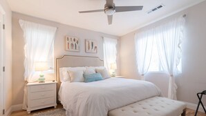 Quarto com cama, mesas de cabeceira, um banquinho e ventilador de teto.