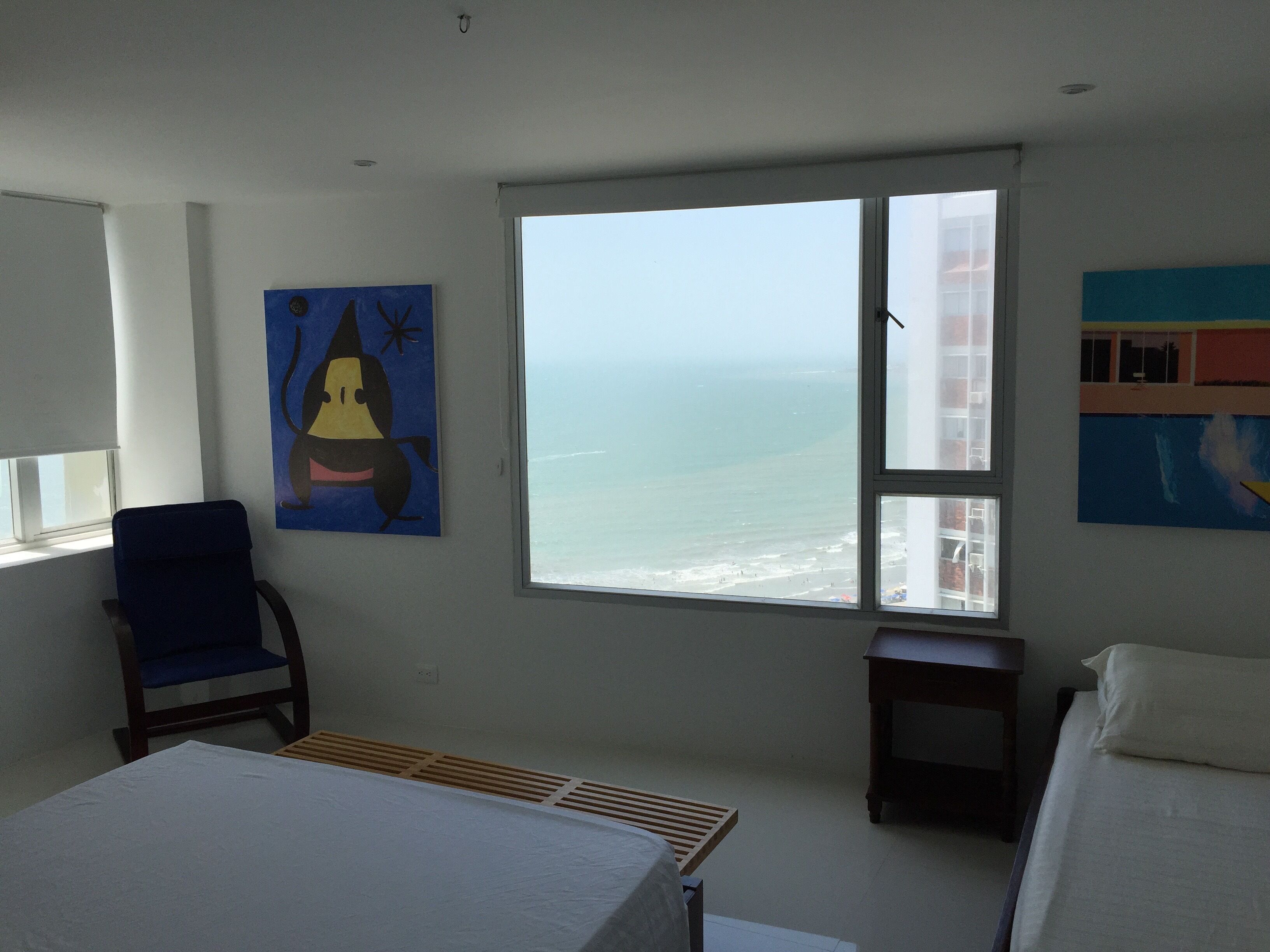 A bedroom with a large window, a bed, a chair, and artwork on the walls.