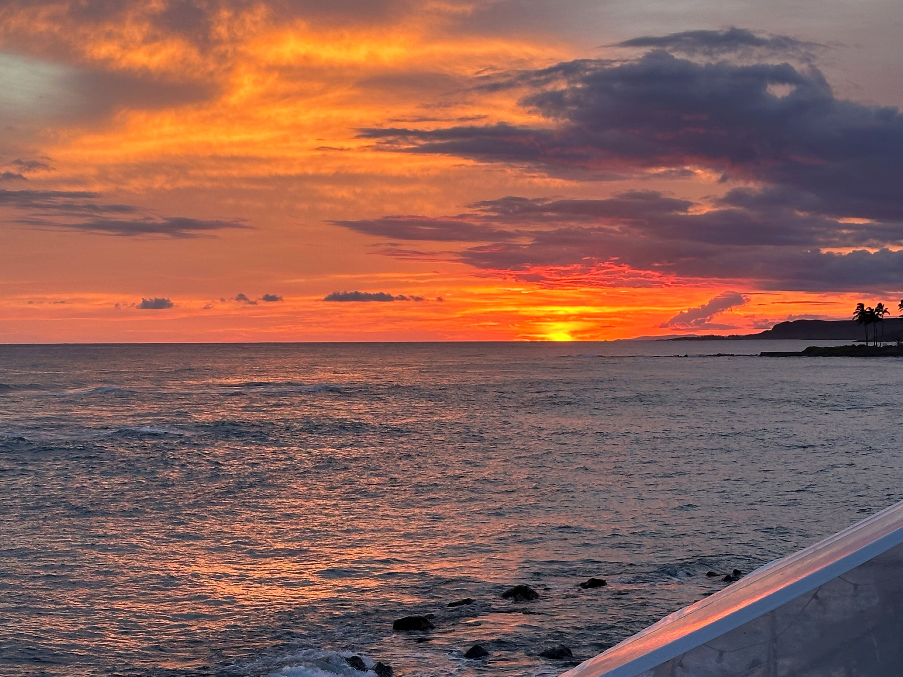 Pôr do sol sobre o oceano, com uma praia e pedras em primeiro plano.