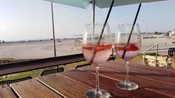 Two glasses of a pink drink with straws on a wooden table with a view of the beach.