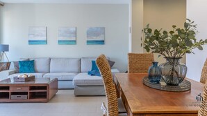 Ein modernes Wohnzimmer mit einer Couch, einem Couchtisch und einem Esstisch. An der Wand hängen drei Gemälde, auf dem Esstisch steht eine Pflanze und links eine Lampe.