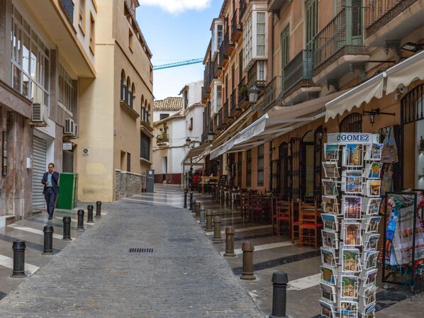 Una calle estrecha flanqueada por edificios, un hombre caminando y un quiosco con postales.