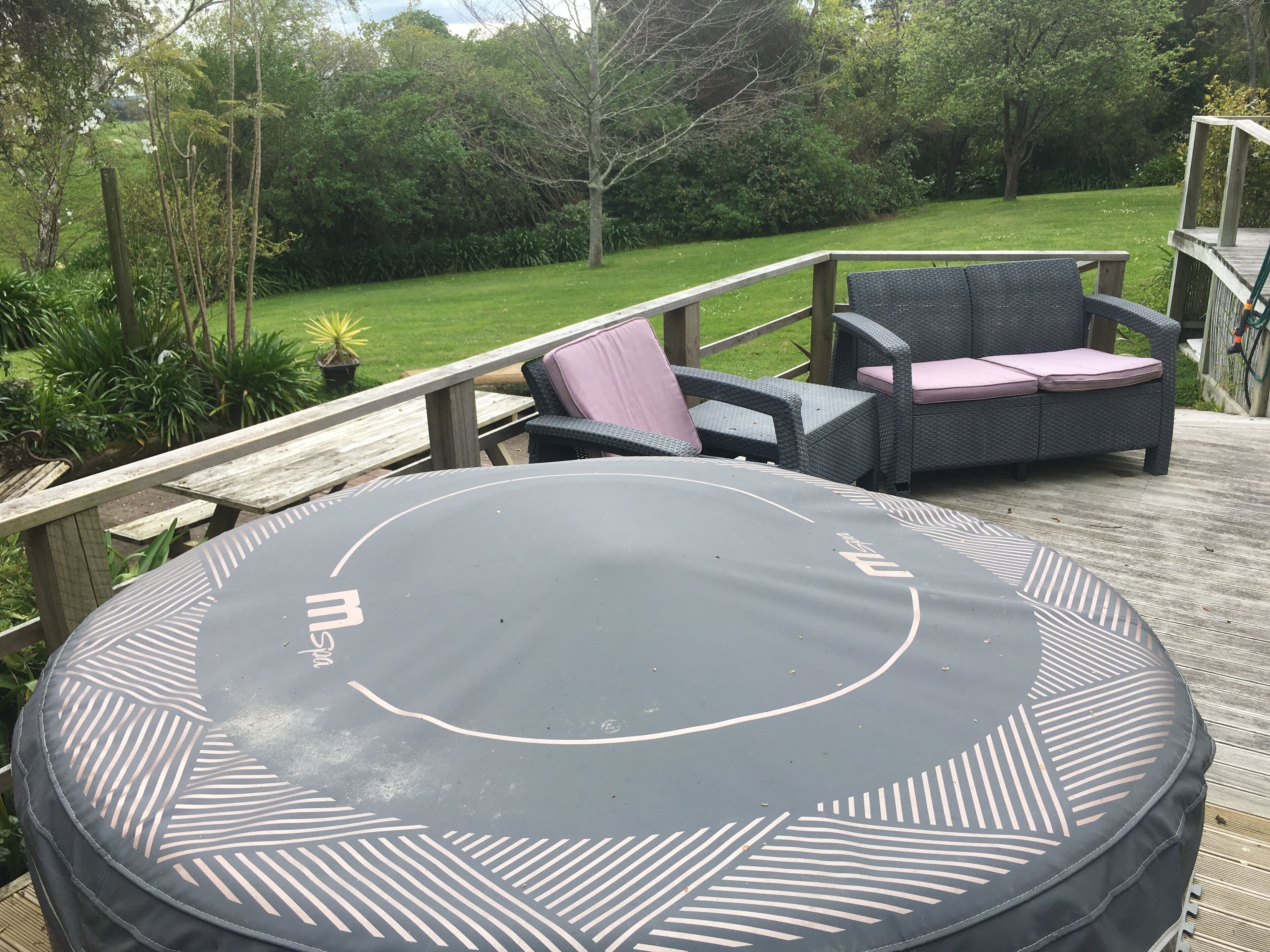 A circular trampoline with a logo, surrounded by wicker chairs with cushions, on a wooden deck with a view of a grassy area and trees.