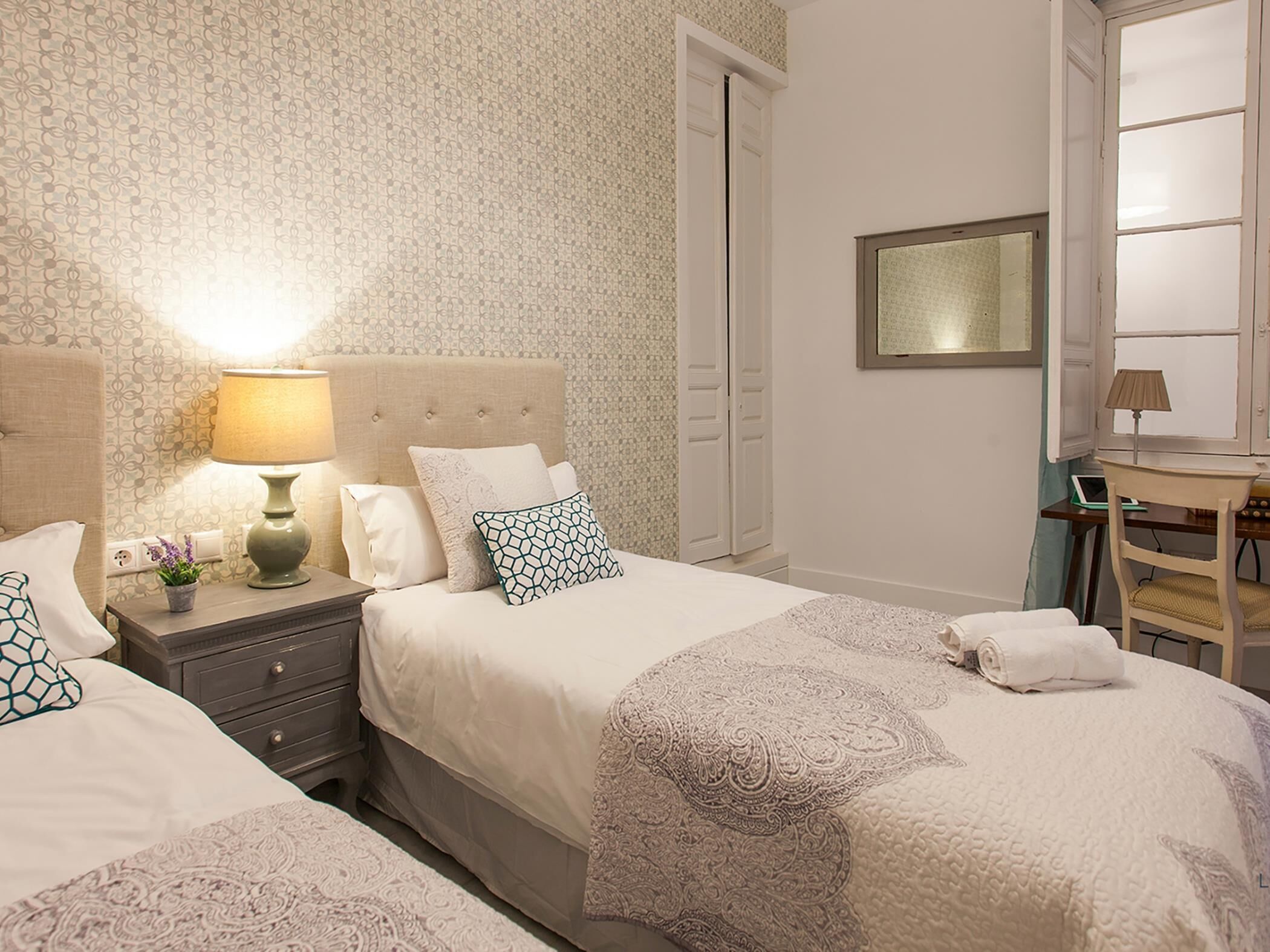 A hotel room with two beds, a nightstand with a lamp, a desk with a laptop, and a large mirror on the wall.