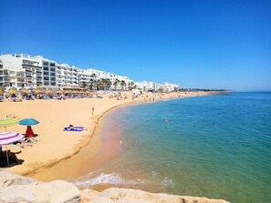 Une plage bordée de bâtiments blancs, avec une eau d’un bleu limpide, et des gens qui se prélassent au soleil.
