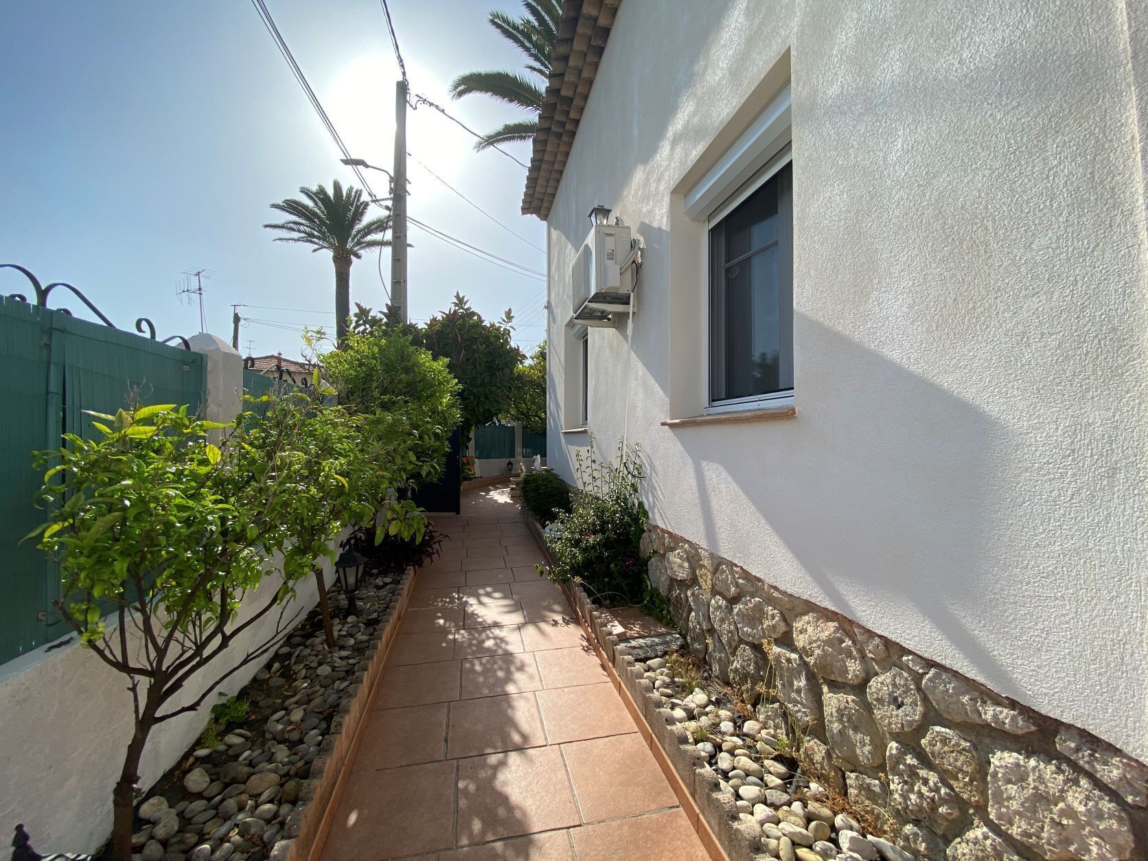 A narrow pathway flanked by a white building and a green fence, leading to a house with a stone wall and a window.