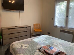 Ein Schlafzimmer mit Bett, Schminktisch, einem orangefarbenen Sessel, einem an der Wand befestigten Fernseher und einem Fenster mit Blick auf Bäume.