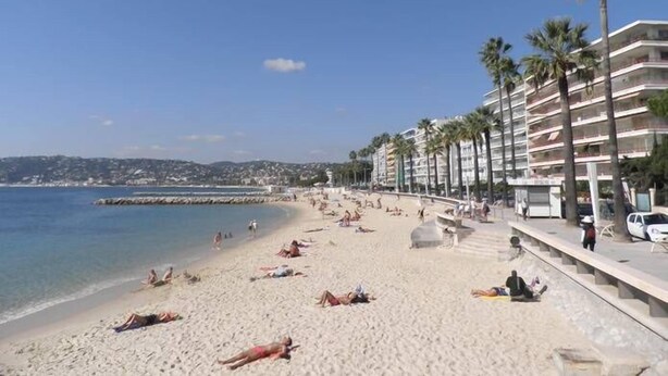 Une plage où des gens se prélassent au soleil, un ciel dégagé et une rangée de palmiers et de bâtiments qui bordent le rivage.