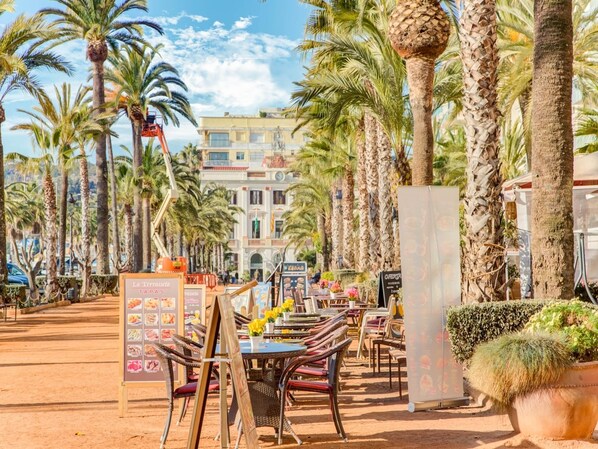 Un área de comedor al aire libre con sillas y mesas de mimbre, rodeada de palmeras y un edificio al fondo.