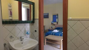A bathroom with a sink, mirror, and toiletries. A bed with a blue bedspread and pillows is visible in the background. - In the center of Agrigento (Agrigento)