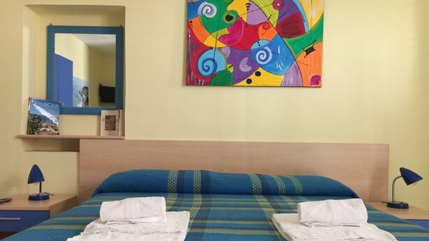 A hotel room with a bed, two pillows, a colorful abstract painting, a mirror, and a nightstand with a lamp. - In the center of Agrigento (Agrigento)