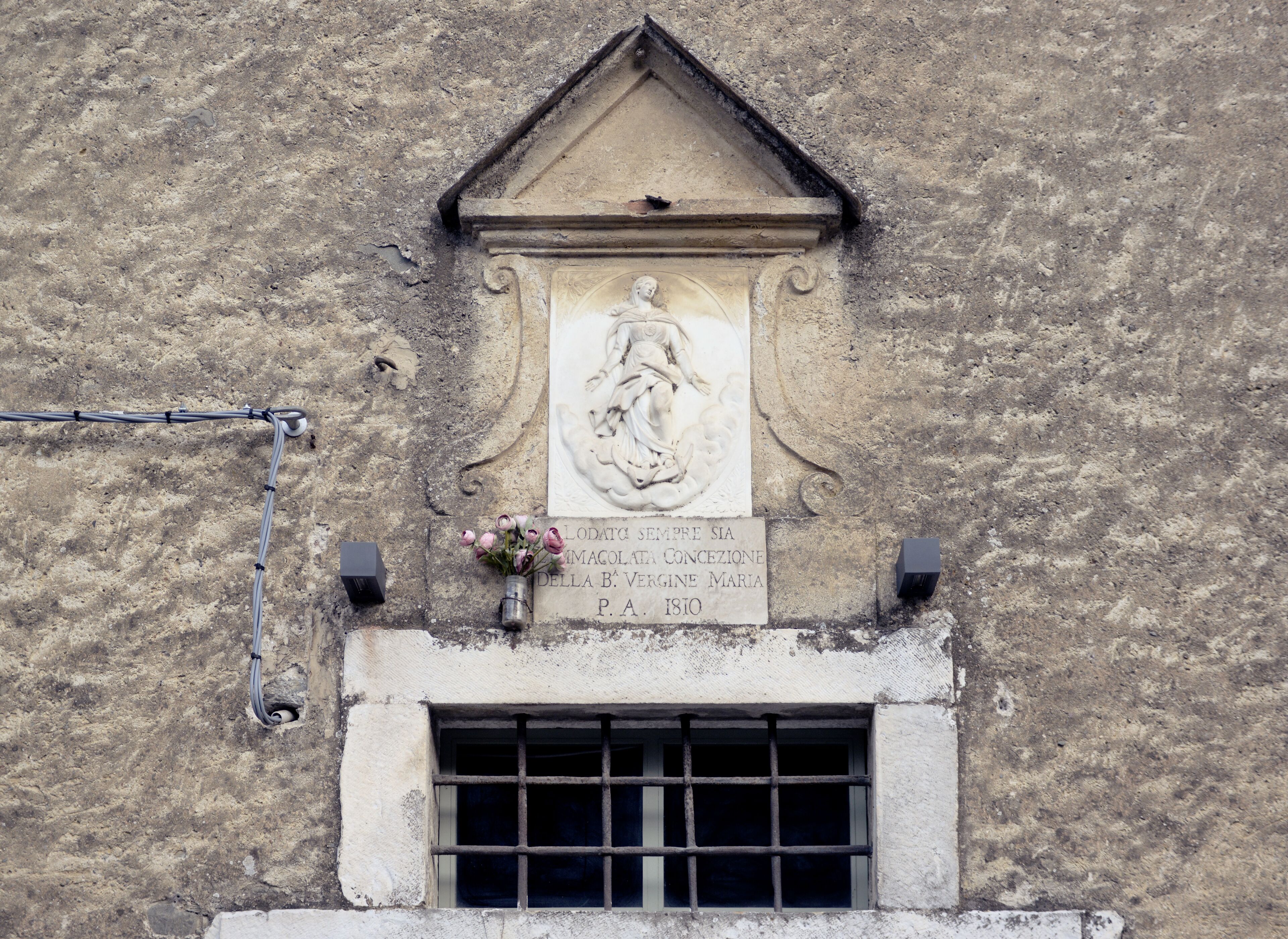 Fachada de un edificio desgastado con una escultura en relieve de piedra sobre una ventana, una placa con texto y un pequeño florero con flores.