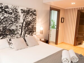 Ein Hotelzimmer mit Bett, Nachttisch, Wandlampe, einer Tür zu einem Badezimmer und Blick auf eine Pflanze draußen.