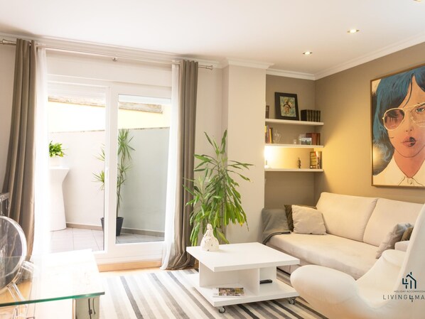 A modern living room with a sofa, coffee table, and a bookshelf. There is a sliding glass door leading to an outdoor area with plants.