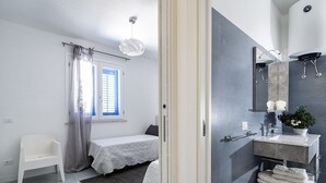 Quarto moderno com cama, uma cadeira branca pequena, janela com cortinas e banheiro com pia, espelho e porta-toalhas.