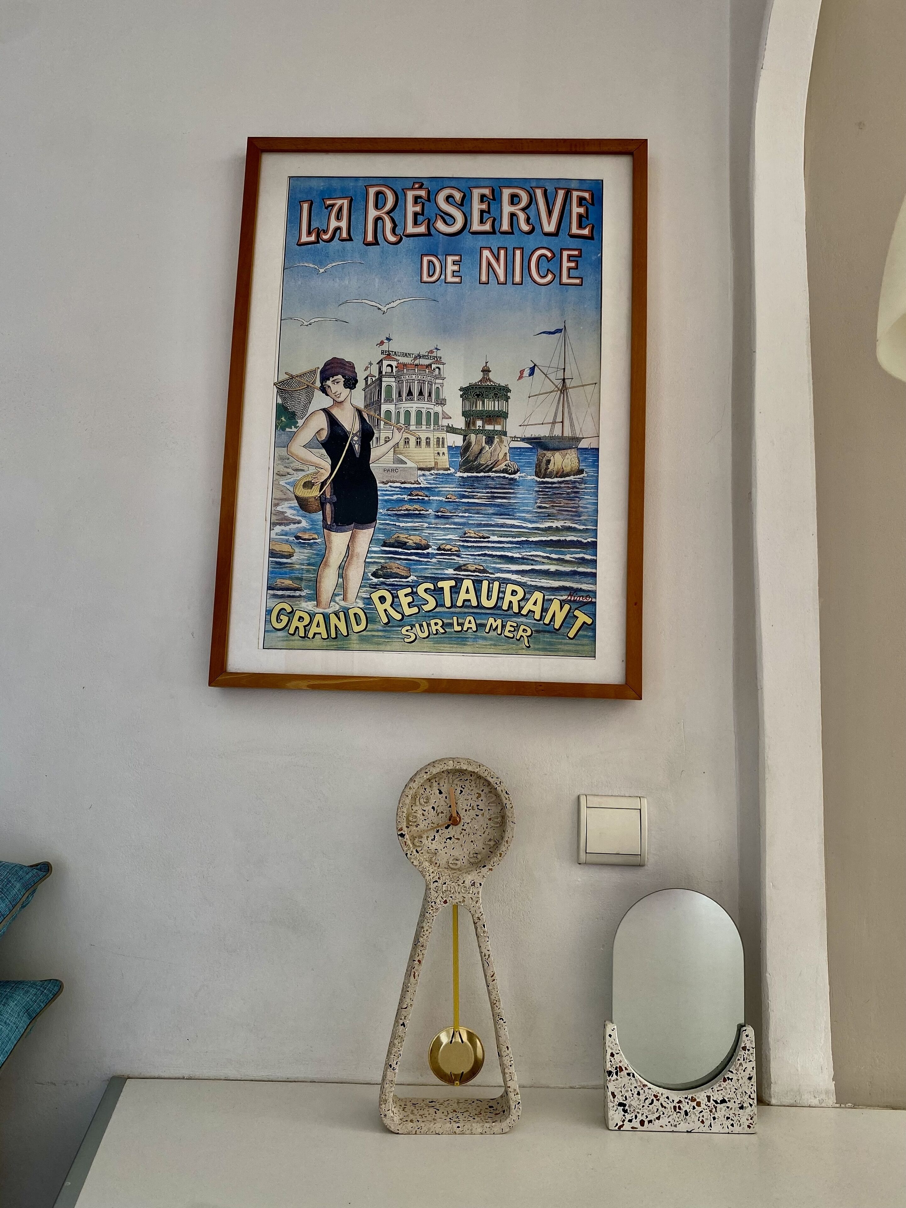 A framed poster of 'La Reserve de Nice' on a wall, a decorative metal object, and a small mirror.
