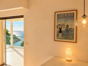 A room with a view of the sea, a framed poster of 'La Reserve de Nice,' and a table lamp.