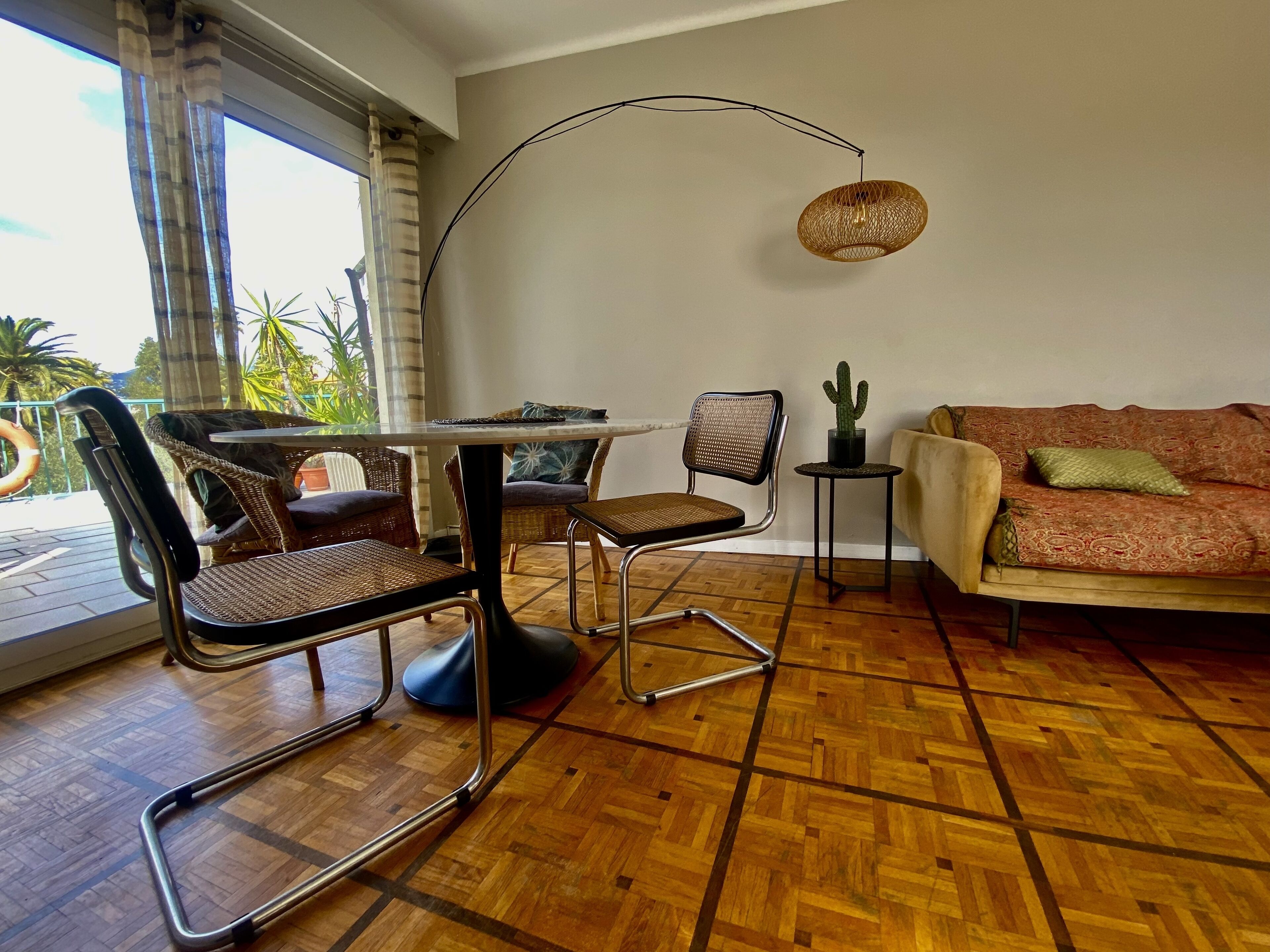 Una stanza con pavimento in parquet di legno, un tavolo rotondo con sedie, un divano, una lampada a sospensione e una pianta di cactus.