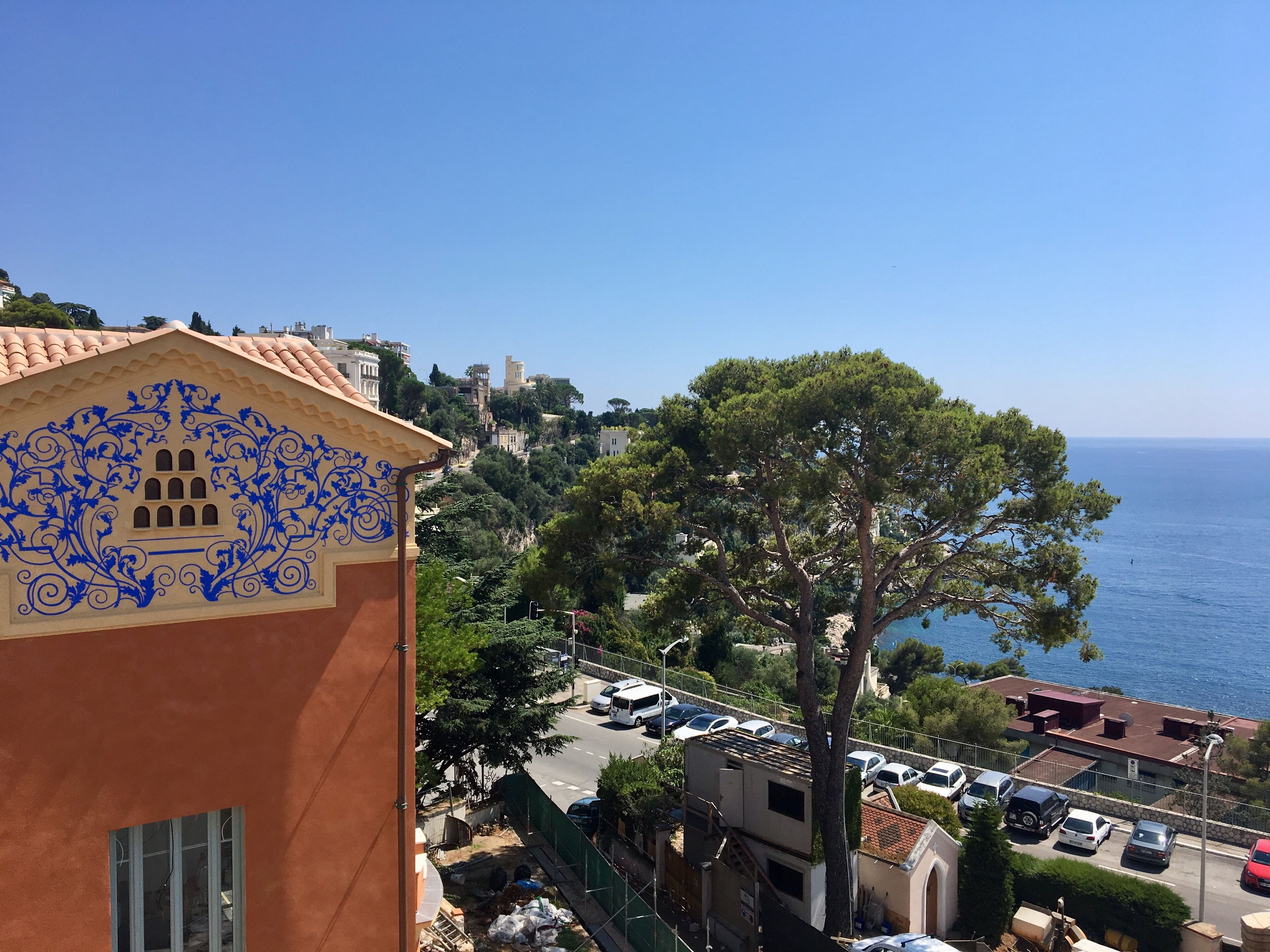 A coastal town with a prominent building featuring blue decorative patterns, a clear view of the sea, and parked cars along the street.