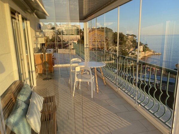 Un balcón con mesa y sillas, un banco de madera y vista al mar y edificios.