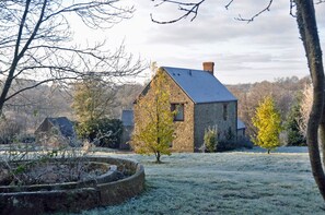 Una casa de piedra con techo inclinado y chimenea, rodeada por un campo cubierto de escarcha y árboles.