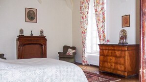 Een slaapkamer met een houten bed, een dressoir, een stoel en een raam met bloemen gordijnen.