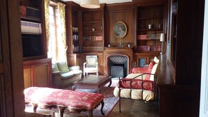 Een klassieke bibliotheek met houten boekenkasten, een open haard en antieke meubels, waaronder een bank, fauteuils en een salontafel.