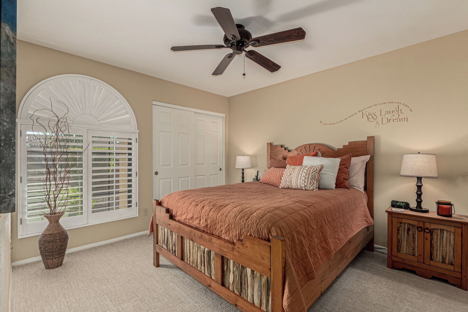A bedroom with a wooden bed, a ceiling fan, a bedside table with a lamp, and a decorative wall piece.