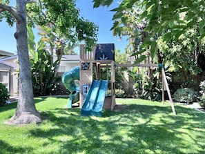 Un jardin avec une structure de jeux en bois, comprenant un toboggan et une zone d’escalade, le tout entouré d’arbres et d’herbe.