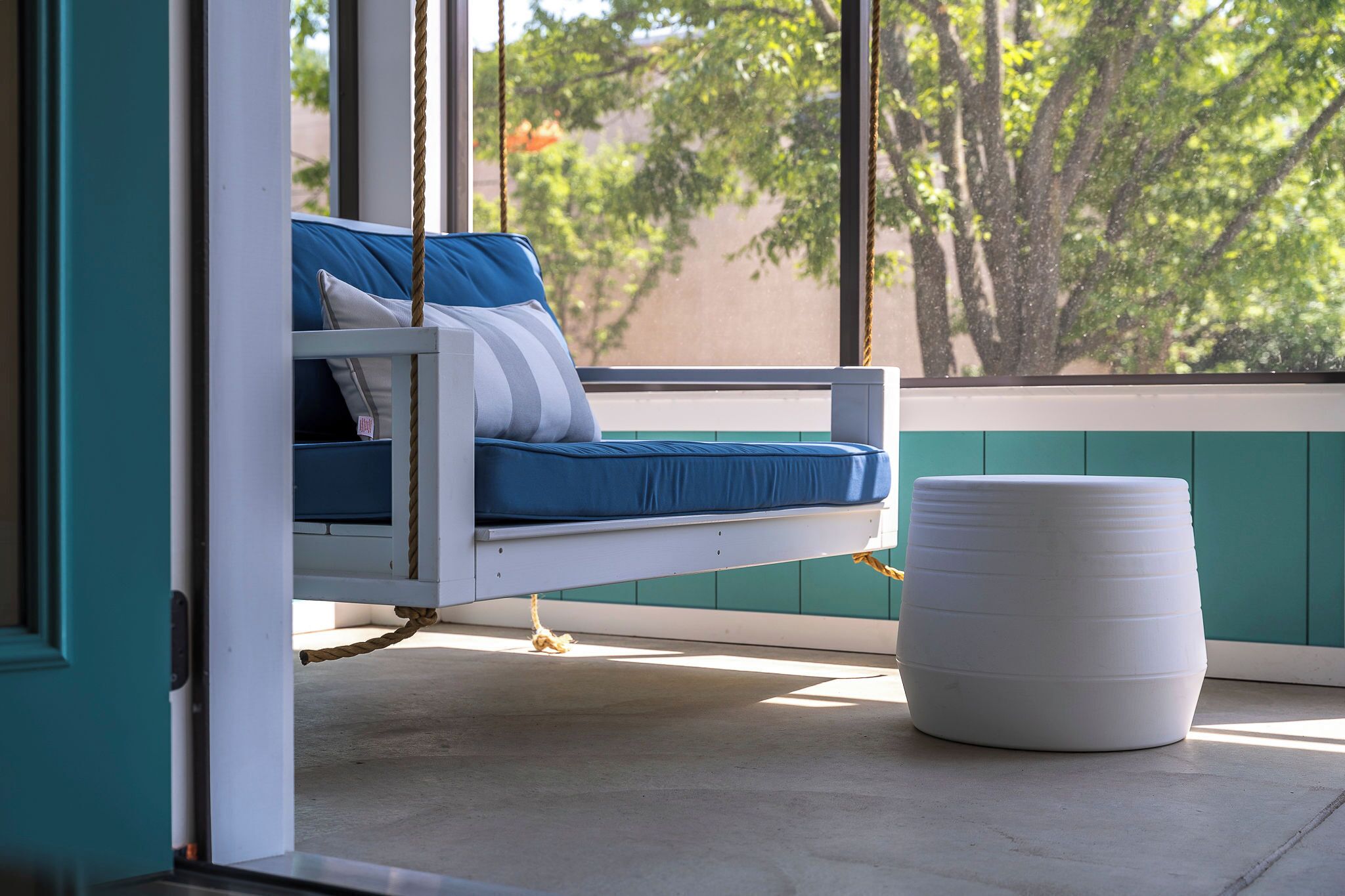 Un lit suspendu avec un matelas bleu et des oreillers blancs, un pouf blanc et une vue sur les arbres à travers de grandes fenêtres.