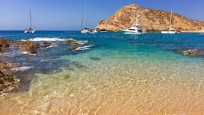 Uma paisagem costeira com águas turquesa cristalinas, margem rochosa e barcos ancorados perto de uma pequena ilha rochosa.