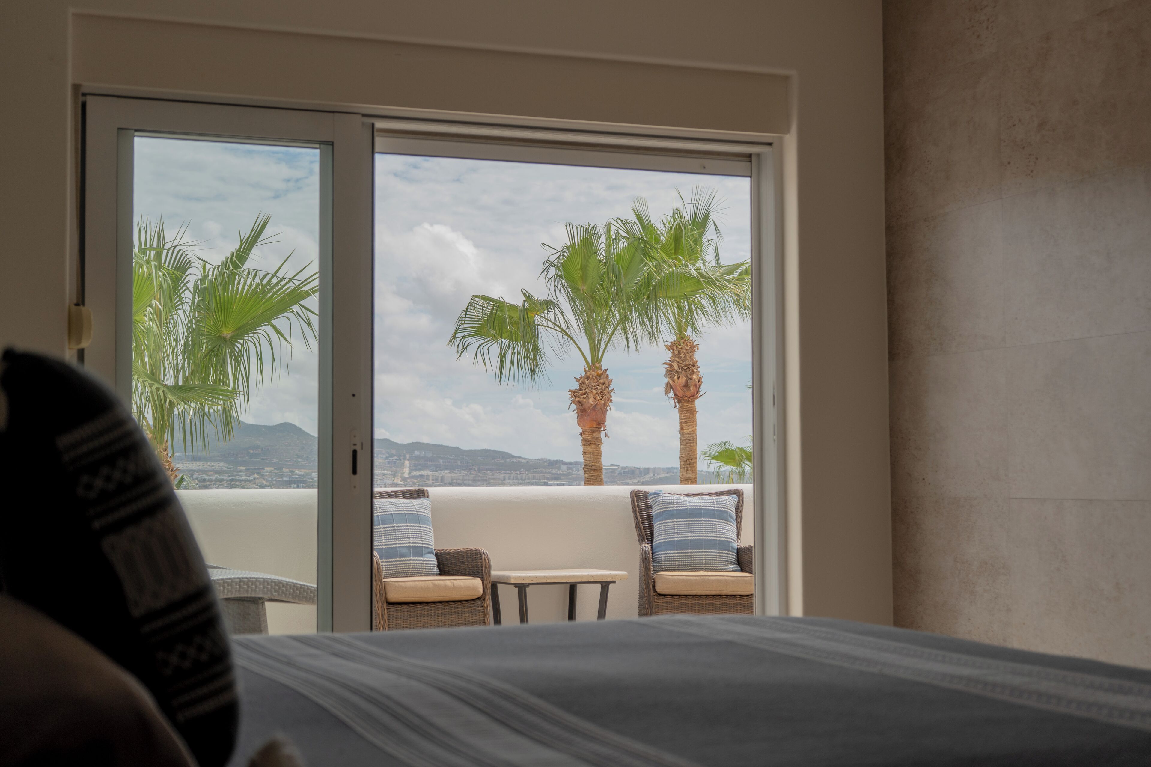 Quarto de hotel com cama, duas cadeiras de vime, uma mesinha e uma porta de vidro deslizante para uma varanda com palmeiras e vista para a cidade.