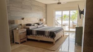 Um quarto espaçoso com uma cama grande, cabeceira de madeira, mesa de cabeceira e vista para o exterior por uma porta de correr de vidro.